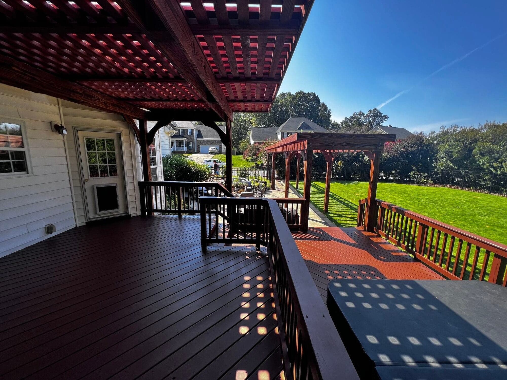 A dark-stained deck running the length of a full cedar lattice pergola, dappled sunlight across the boards with green lawn beyond, built by Berg Custom Builds in the Charlotte metro